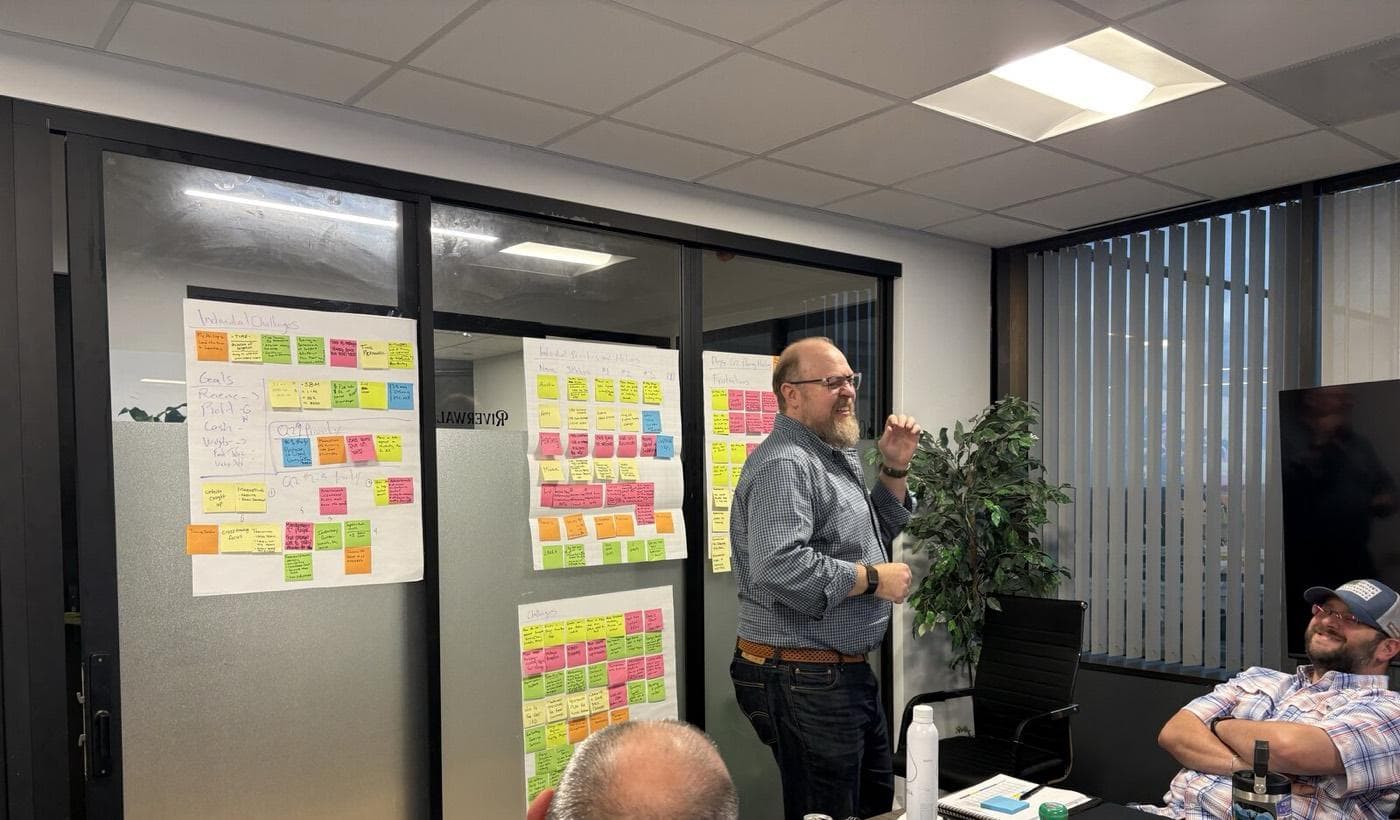 A man stands beside glass walls covered in rows of colored sticky notes while people sit around a conference table with notebooks and drinks.