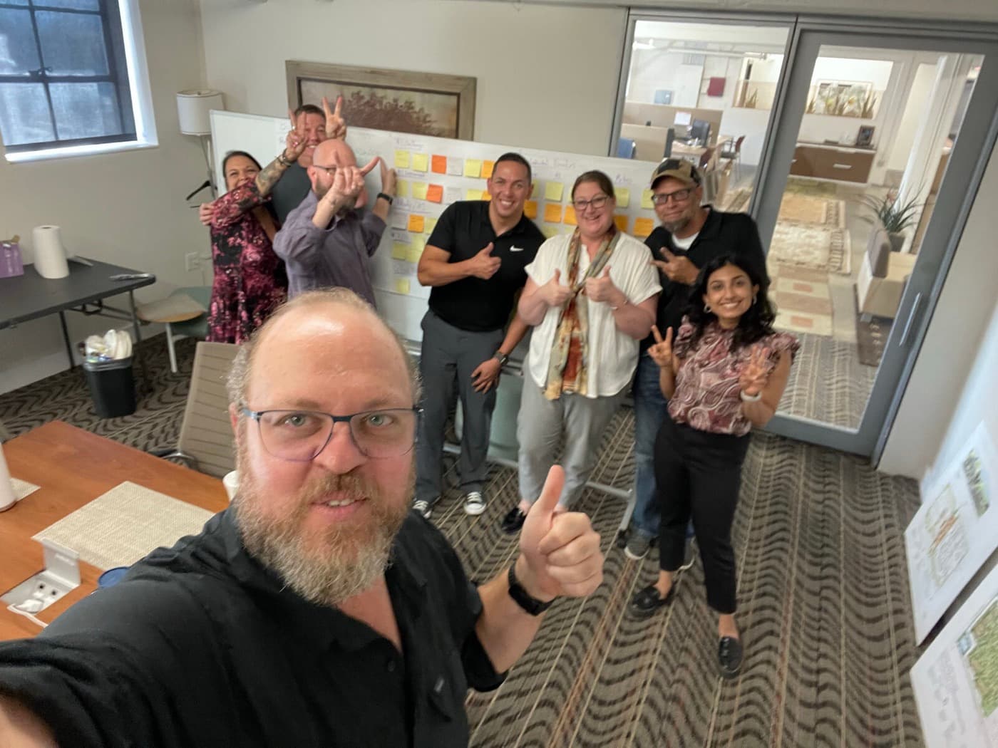 A bearded man in glasses takes a selfie with a thumbs up while six people stand behind him, smiling, in front of a wall covered in colored sticky notes.