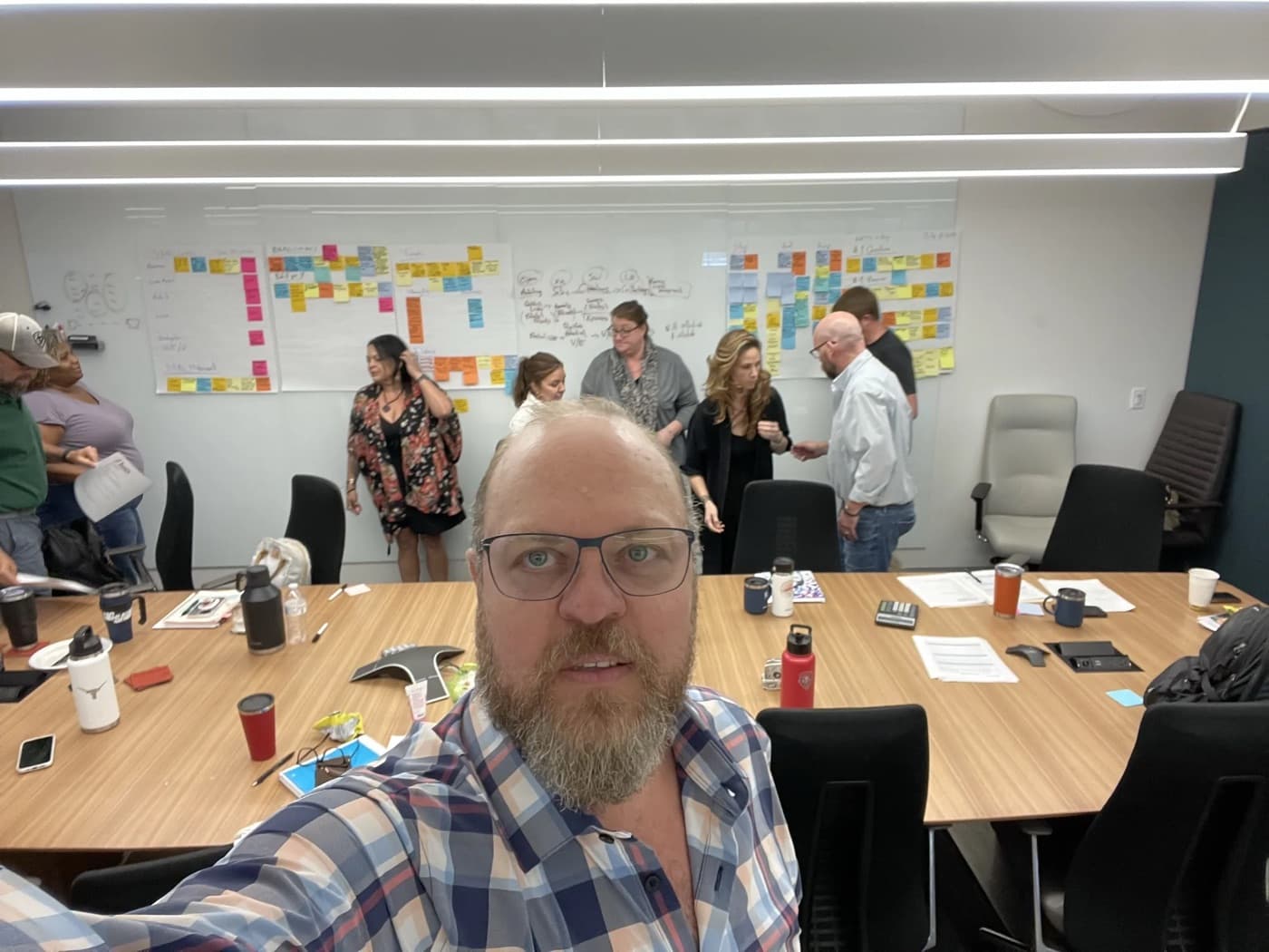 Max Koz takes a selfie in a client working session. Behind him, the owner's team stands at a long wall covered in colored sticky notes and flip-chart sheets.